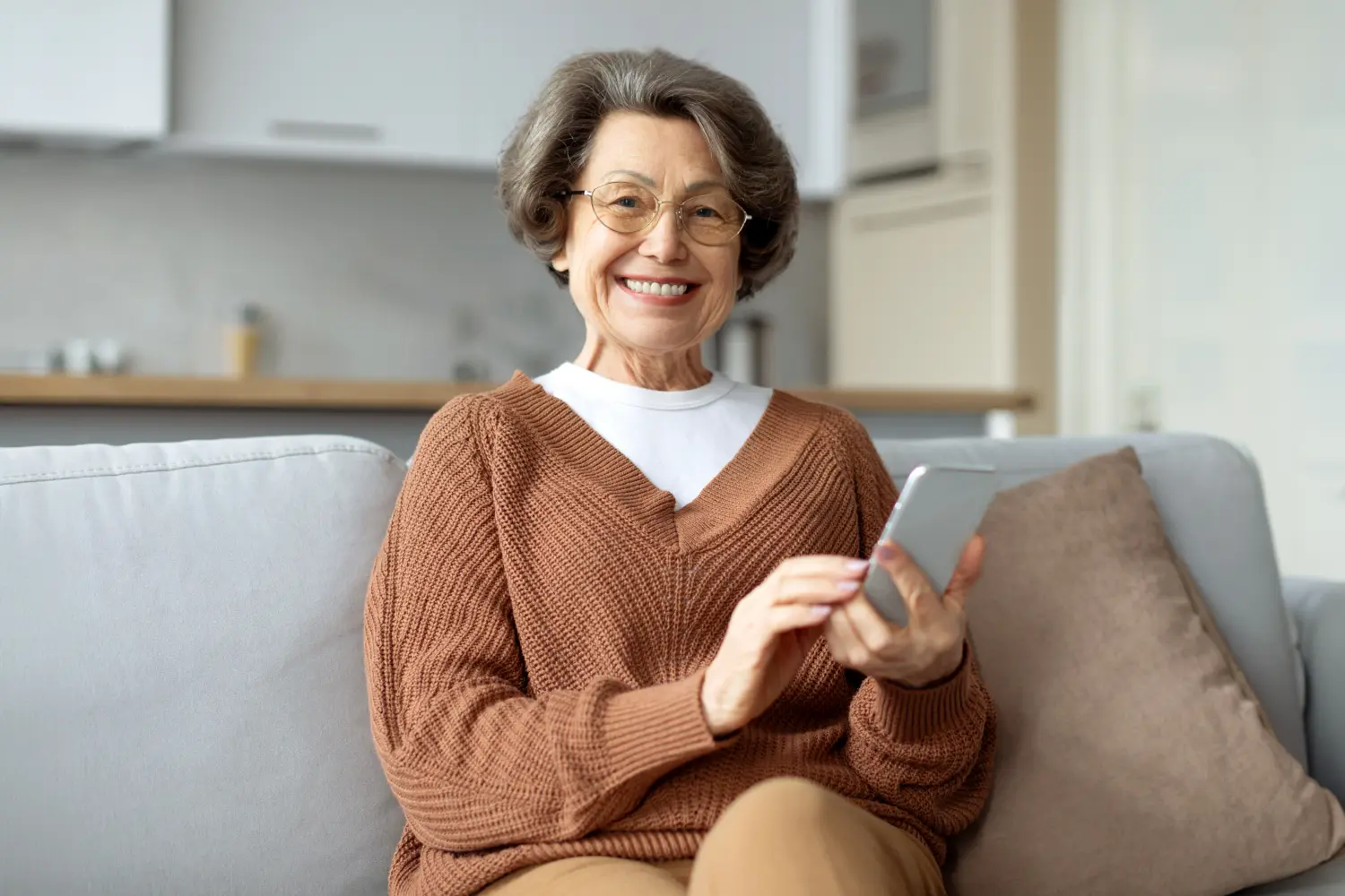 Rentnerin, die eine Nachricht auf ihrem Handy liest und sich über eine gute Nachricht freut, lächelt in die Kamera und genießt die drahtlose Verbindung auf der heimischen Couch